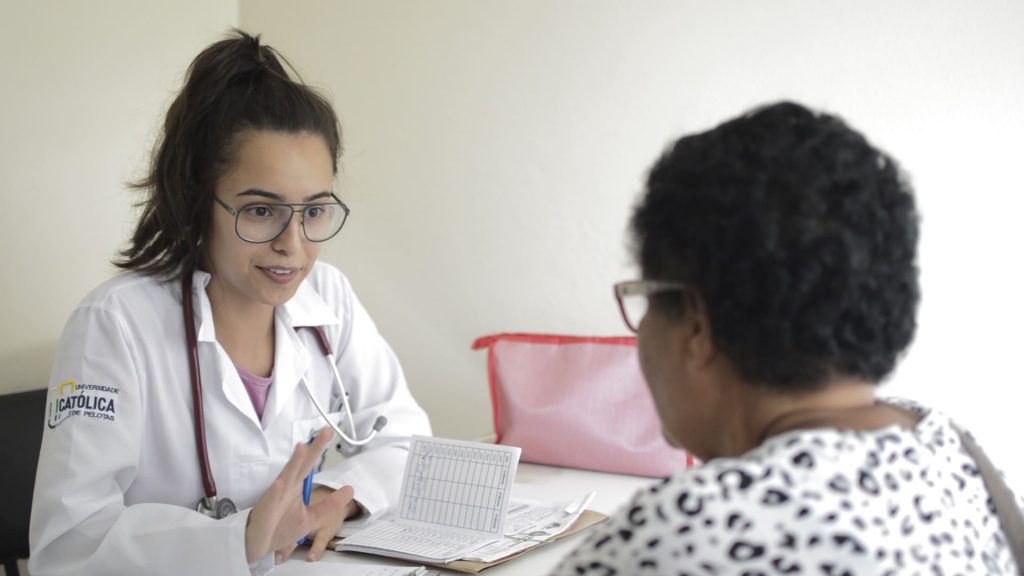 Uma aluna, da Medicina da UCPel, branca, de jaleco e óculos, caneta na mão, sentada em uma mesa, atende uma paciente negra.