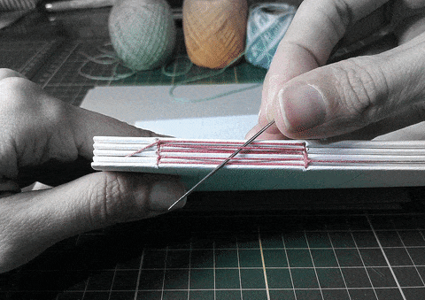 Duas mãos, com uma agulha e linha, costuram folhas de papel.
