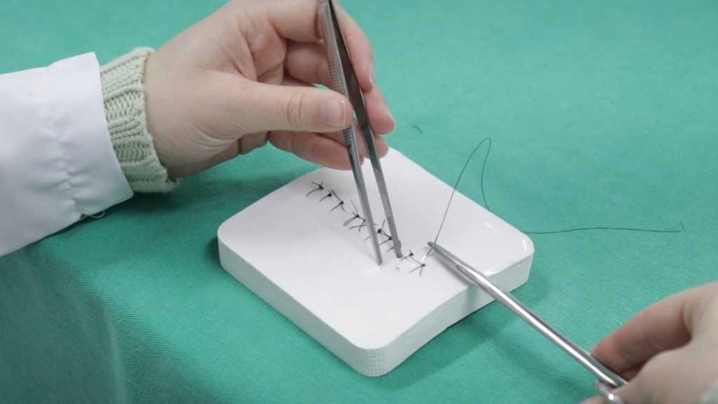 Em close mão com uma pinça realiza sutura em um simulador branco disposto sobre um fundo verde
